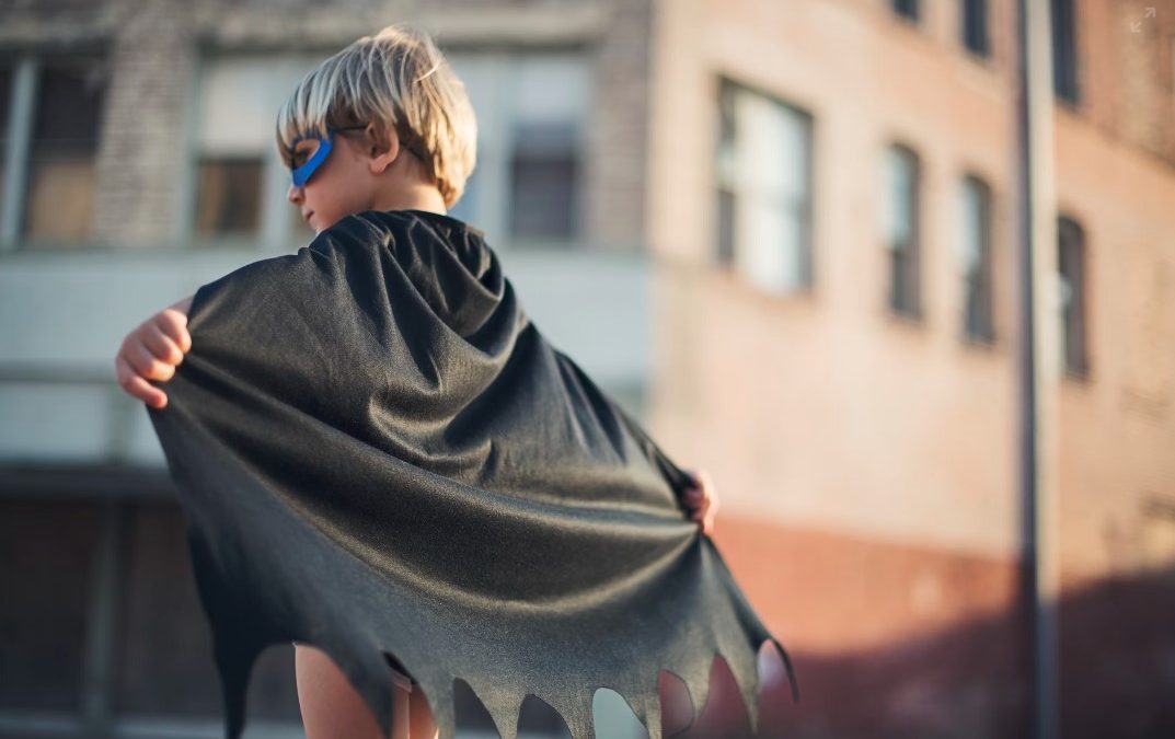 Mazs zēns supervaroņa kostīmā stāv uz ielas izplestām rokām.
